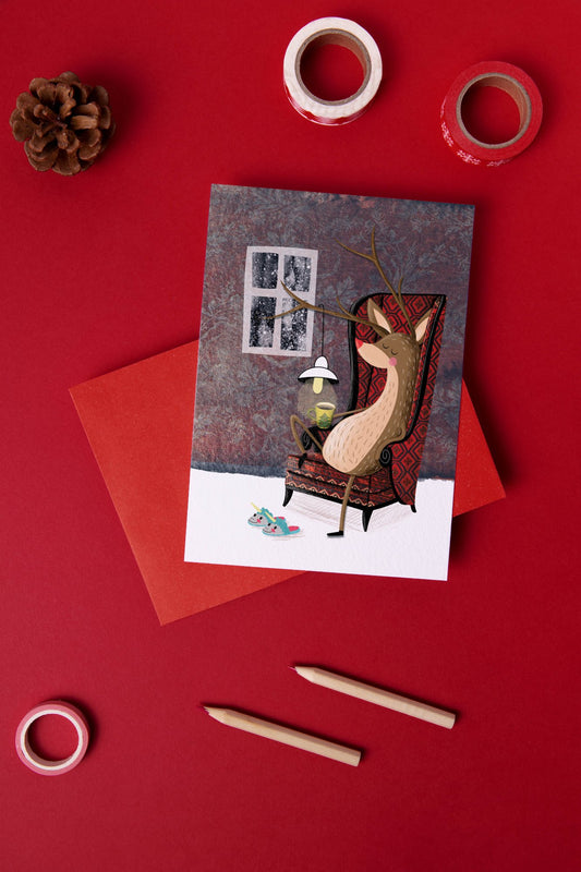 Greeting card featuring a cozy reindeer sitting in a chair with a cup of cocoa and wearing unicorn slippers, perfect for Christmas with reindeer-themed cards.